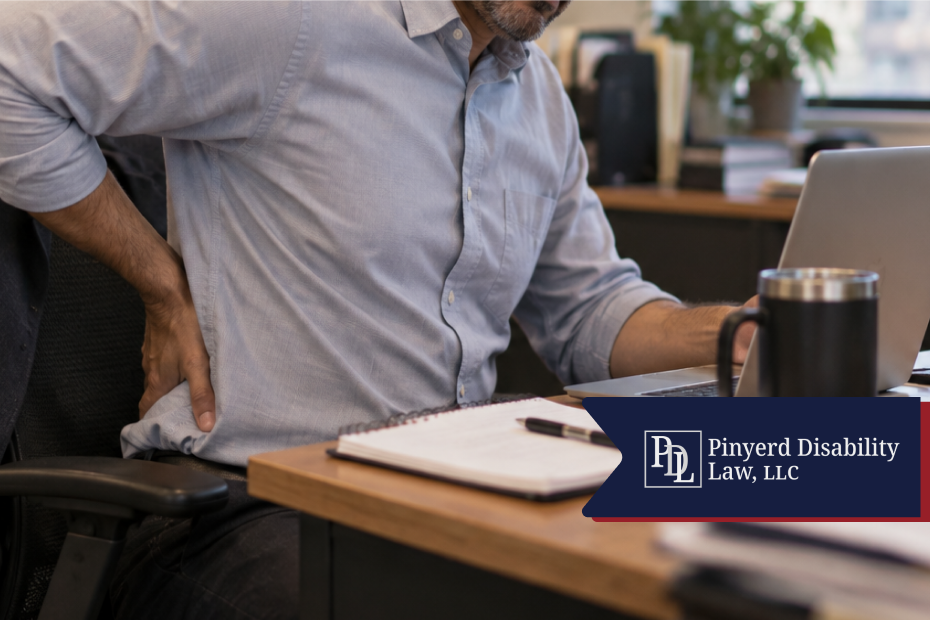 Man at his desk holding his lower back while working on a laptop